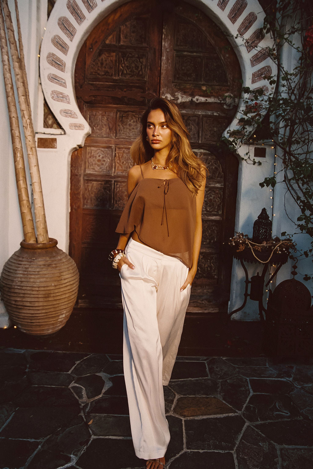 Model wearing the 12th Tribe Moonlit Kiss Cream Satin Trousers, styled with the Costella Mocha Asymmetrical Top, Soreya Resin and Gold Bangles, and Calaia Gold Earrings, featuring a mid rise fit, wide-leg satin silhouette for an elegant, flowing fit