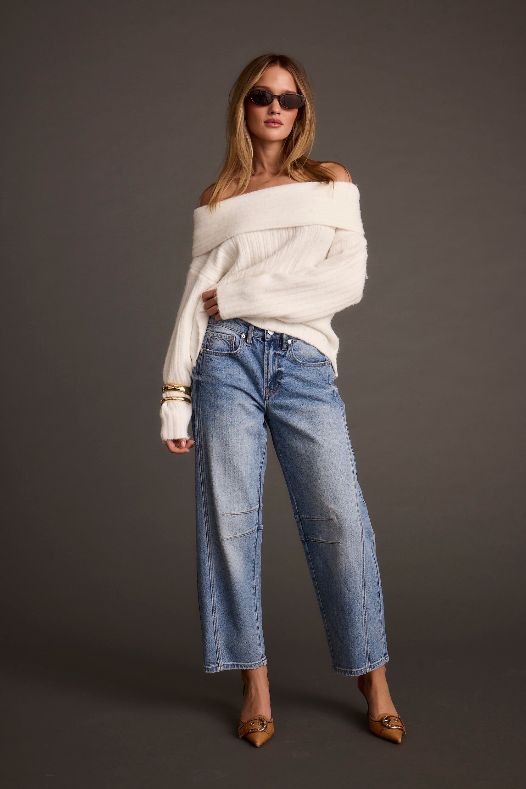 Woman wearing a white off-shoulder sweater and blue jeans against a gray background