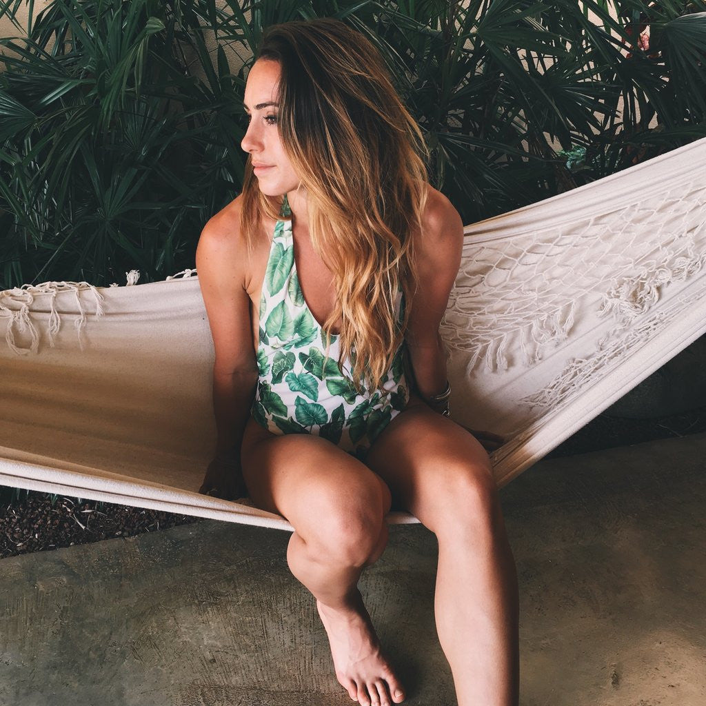 Tropical leaf-print halter swimsuit worn on a fringed hammock beside lush palm foliage.