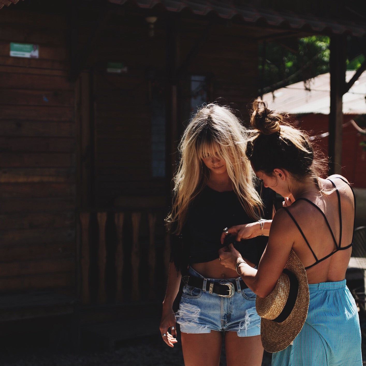 Two women in summer outfits stand outside a wooden cabin, one holding a straw sun hat.