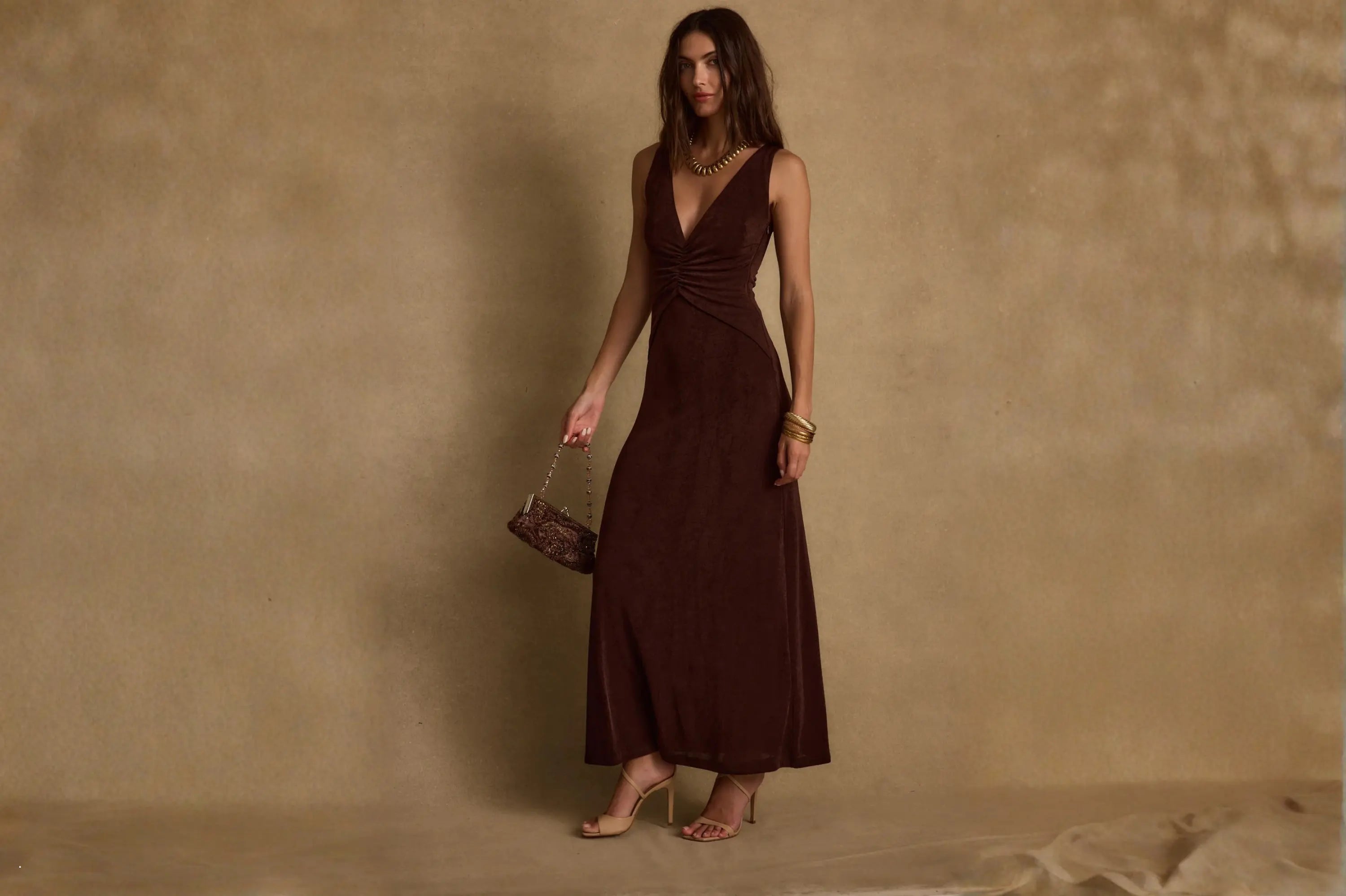 A deep brown v-neck ruched maxi dress styled with gold bangles, strappy heels, and a beaded clutch, a polished wedding guest resort wear look.