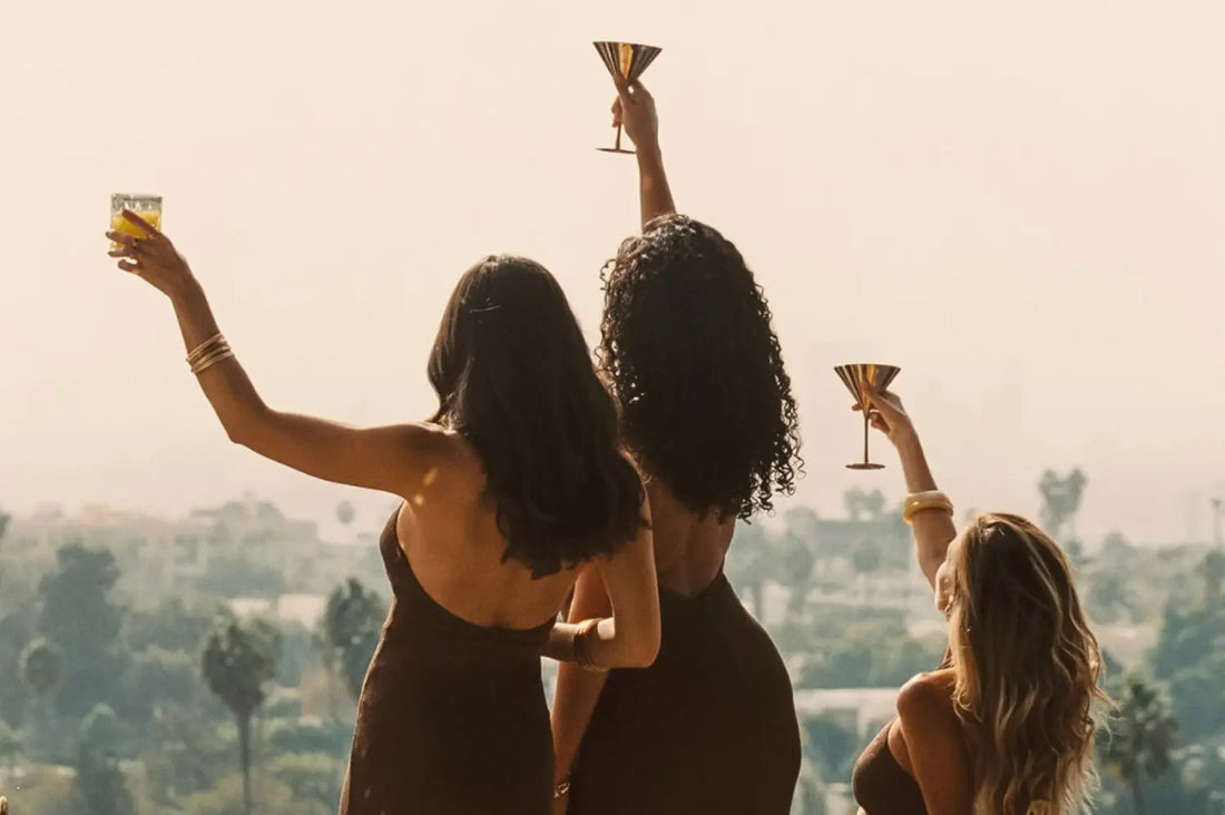 Three friends in elegant resort wear for women toast with cocktails on a sunlit balcony