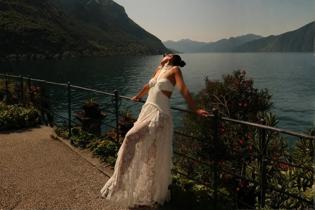 A woman in a white lace crop top and flowing skirt leans against a lakeside railing with mountain views, wearing dreamy summer vacation clothing.