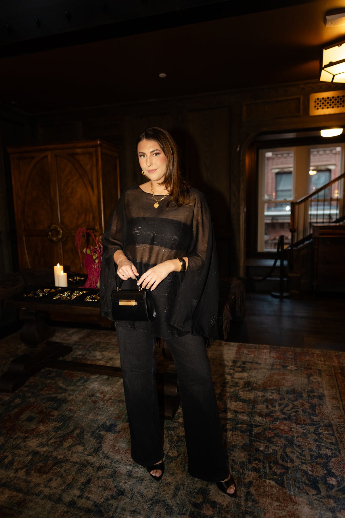 Woman wearing a sheer black long-sleeve top with a horizontal band and black wide-leg pants, standing in a dimly lit indoor setting.
