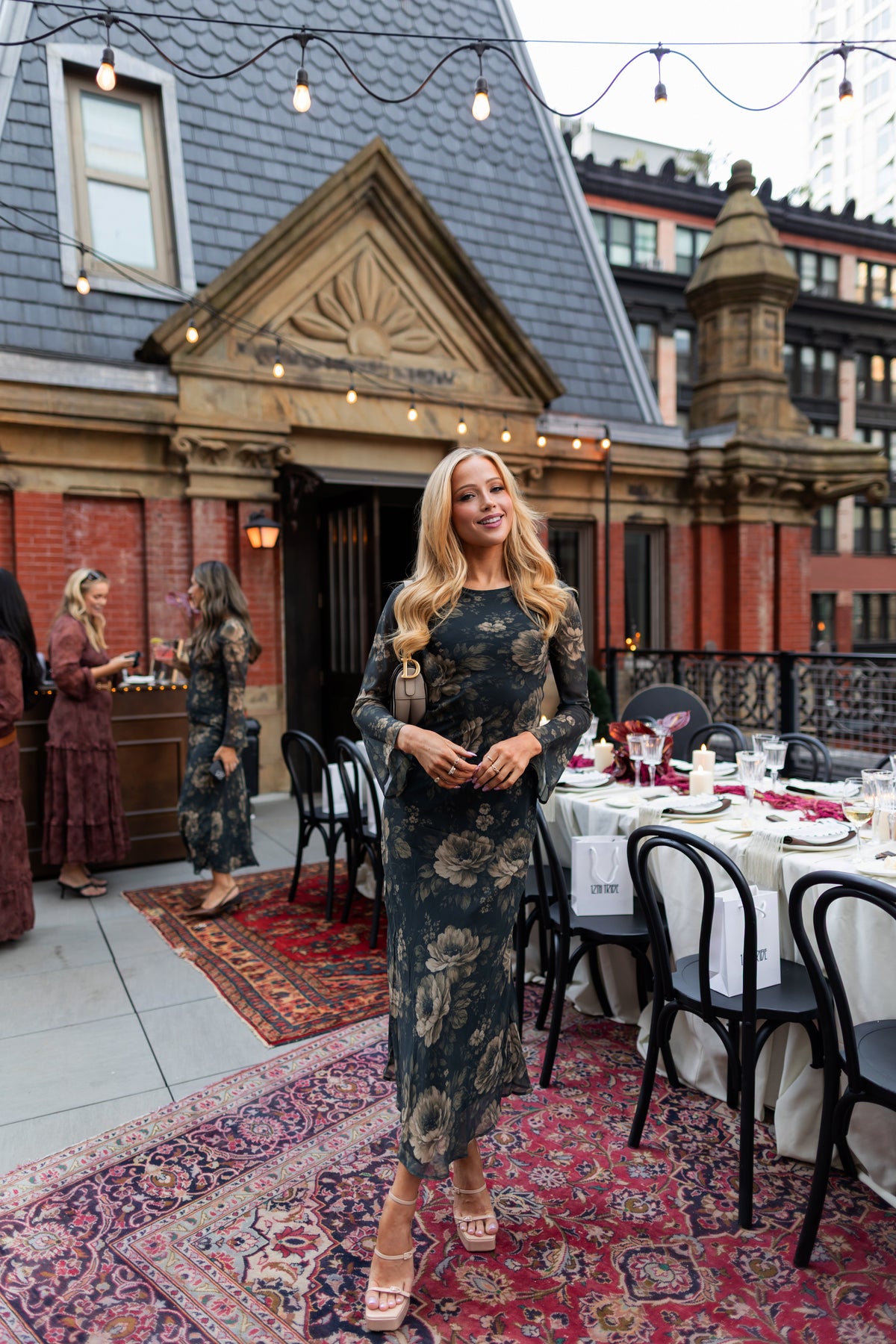 Woman wearing a dark green floral maxi dress with a gold-toned print, standing on a string-lit rooftop patio set for dinner.