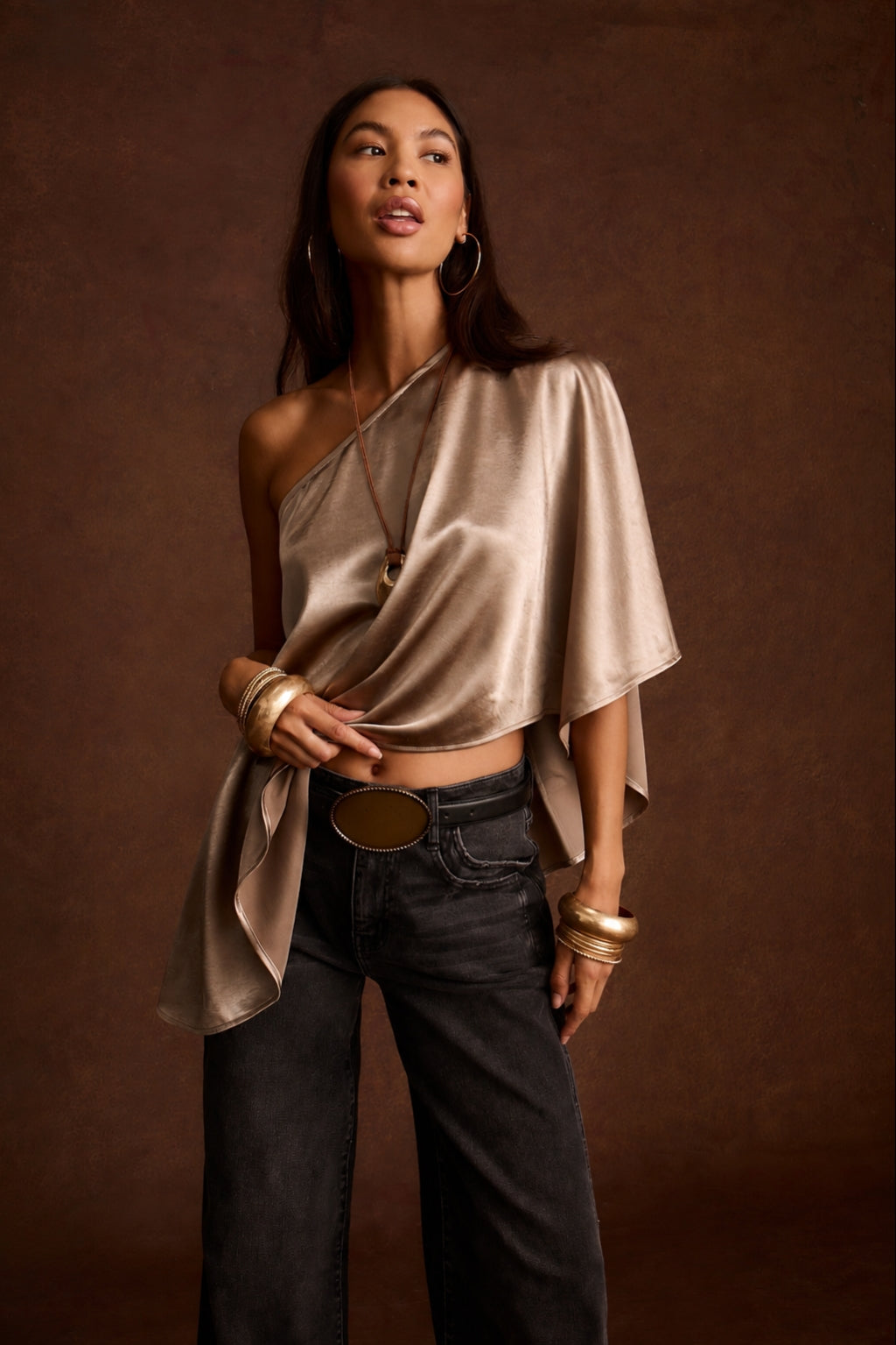 Woman wearing a draped champagne satin one-shoulder top with dark jeans and gold jewelry
