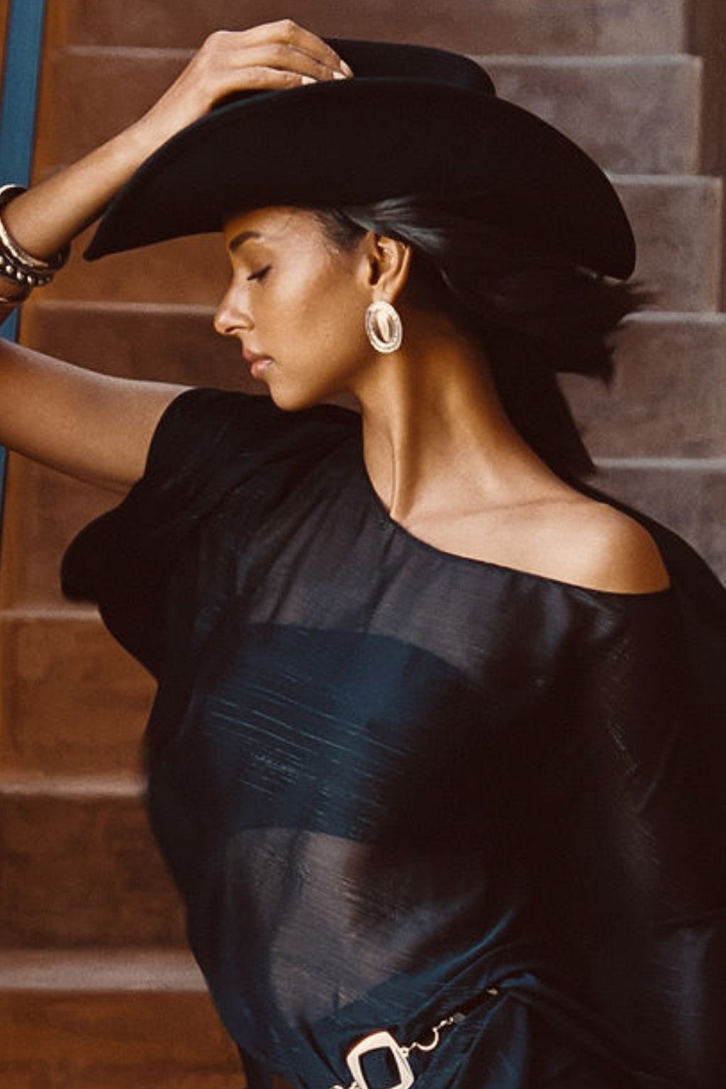 Woman wearing Bailey gold and silver statement hoop earrings with McGraw cowboy hat and black off-shoulder dress on staircase backdrop.