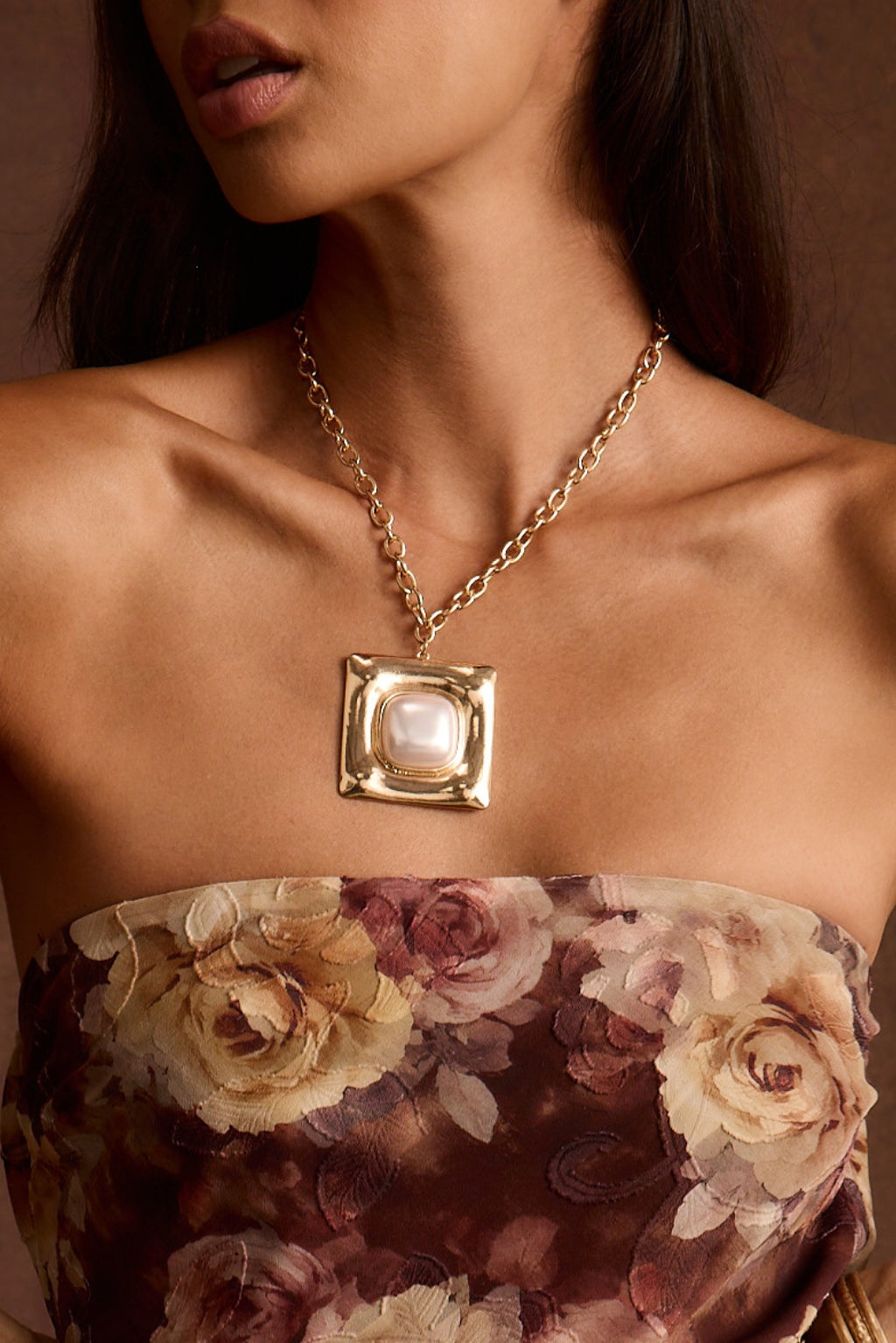 Gold chain necklace with a square pearl pendant worn over a strapless floral top.