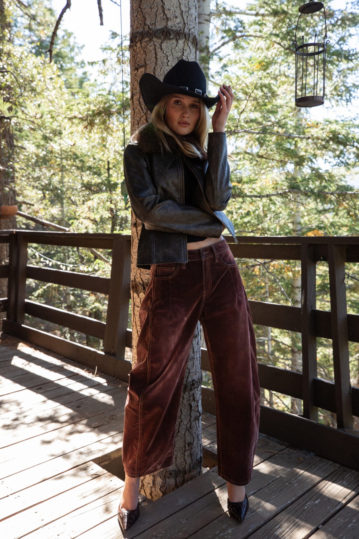 Mulberry velveteen barrel pants styled with a leather jacket and cowboy hat on a wooded deck.