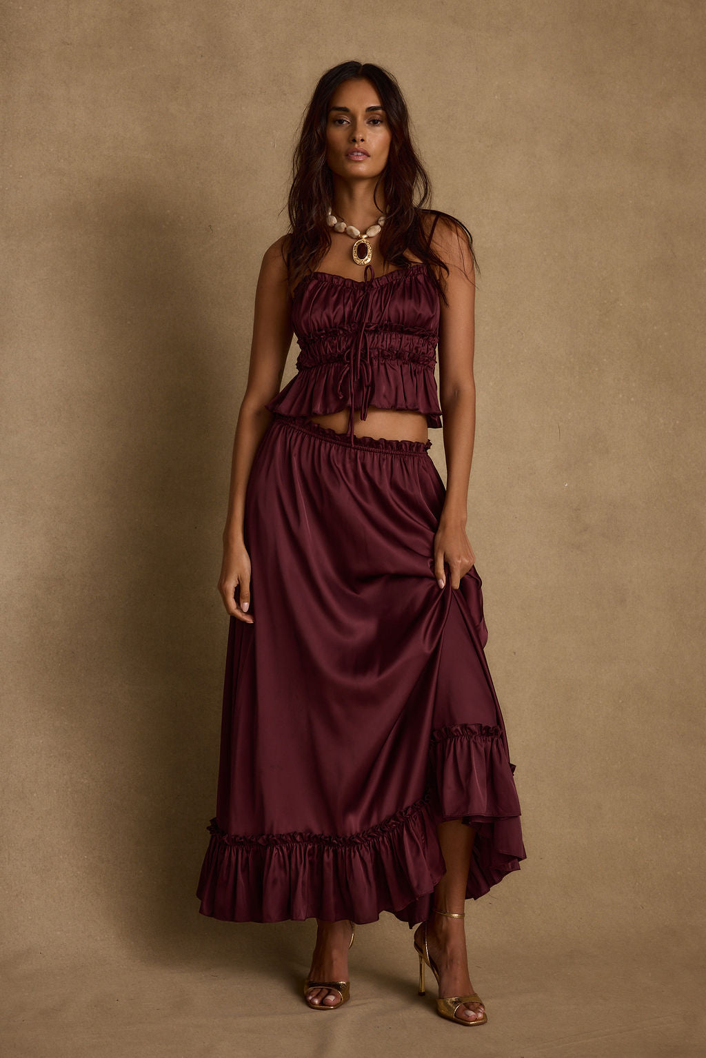 Model wearing the 12th Tribe Dahlia Mulberry Satin Tank and matching Dahlia Mulberry Satin Maxi Skirt in a burgundy satin two-piece set, featuring a front-tie crop top and high-waisted ruffle-hem maxi skirt, styled with strappy heels for bachelorette parties, festivals, and evening events