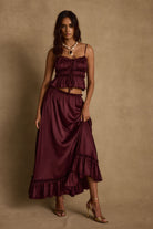 Model wearing the 12th Tribe Dahlia Mulberry Satin Tank and matching Dahlia Mulberry Satin Maxi Skirt in a burgundy satin two-piece set, featuring a front-tie crop top and high-waisted ruffle-hem maxi skirt, styled with strappy heels for bachelorette parties, festivals, and evening events.
