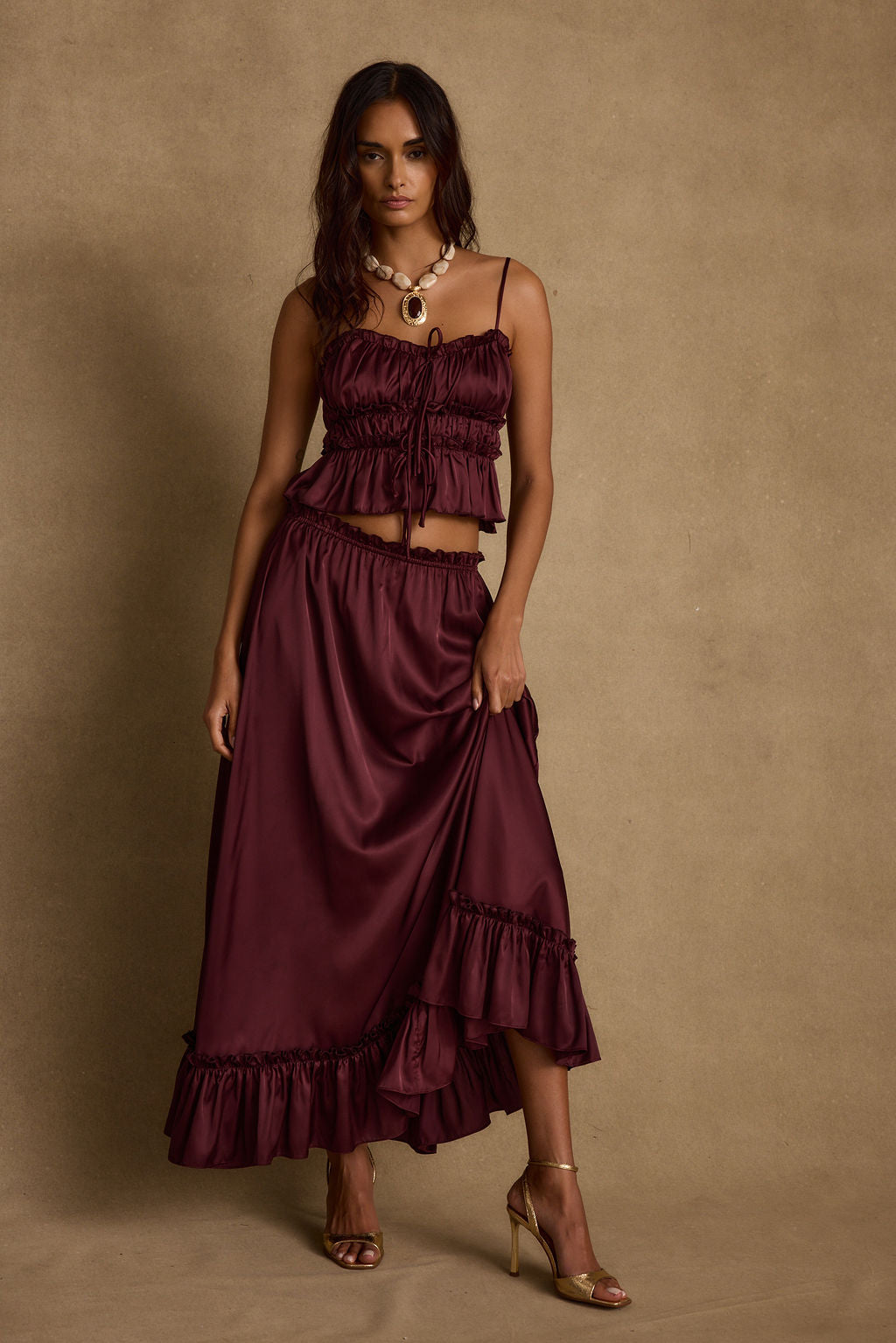 Model wearing the 12th Tribe Dahlia Mulberry Satin Tank and matching Dahlia Mulberry Satin Maxi Skirt in a burgundy satin two-piece set, featuring a front-tie crop top and high-waisted ruffle-hem maxi skirt, styled with strappy heels for bachelorette parties, festivals, and evening events.