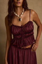 Model wearing the 12th Tribe Dahlia Mulberry Satin Tank and matching Dahlia Mulberry Satin Maxi Skirt in a burgundy satin two-piece set, featuring a front-tie crop top and high-waisted ruffle-hem maxi skirt, styled with strappy heels for bachelorette parties, festivals, and evening events.