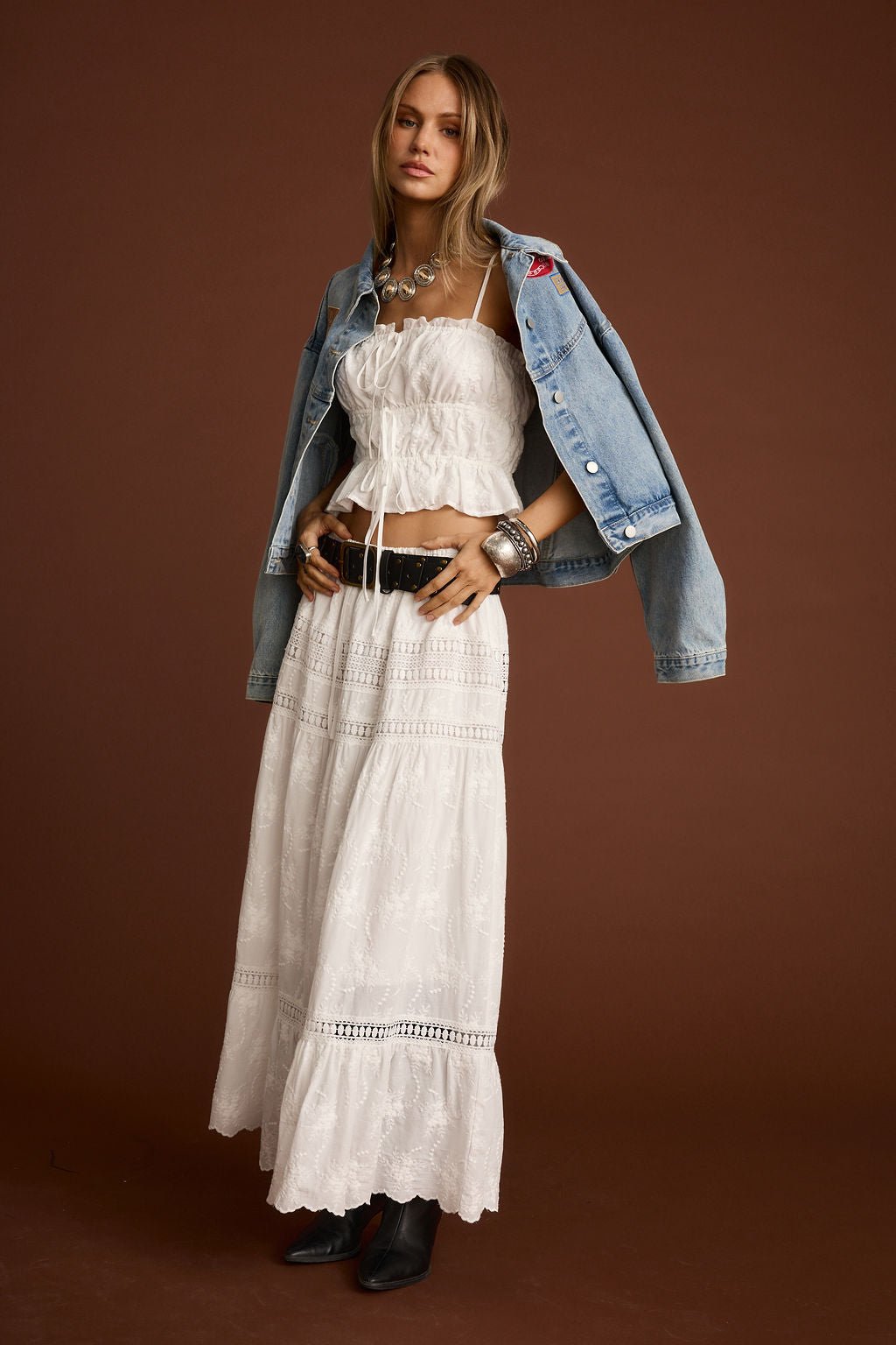 White ruched crop top with eyelet midi skirt, denim jacket, and black studded belt against a brown background