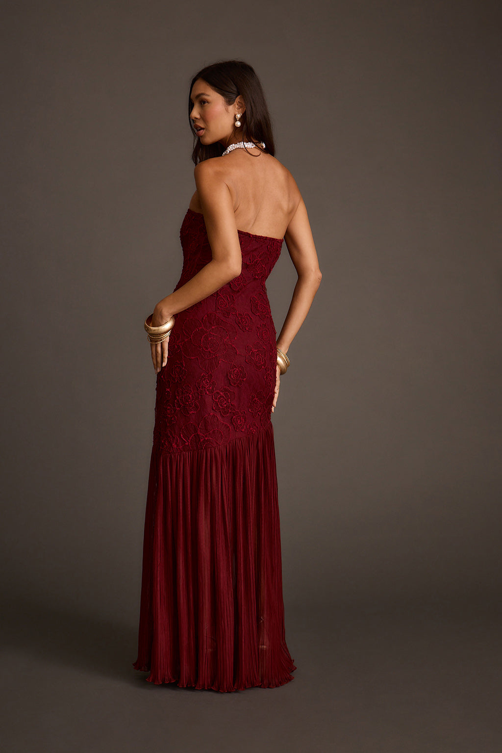 Back view of a strapless burgundy lace maxi dress with a pleated flared skirt.