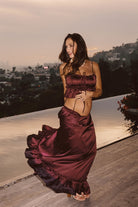 Model wearing the 12th Tribe Dahlia Mulberry Satin Tank and matching Dahlia Mulberry Satin Maxi Skirt in a burgundy satin two-piece set, featuring a front-tie crop top and high-waisted ruffle-hem maxi skirt, styled with strappy heels for bachelorette parties, festivals, and evening events.