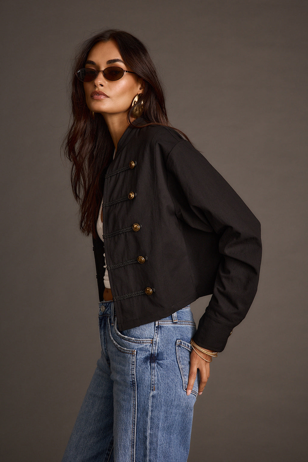 Side view of a woman wearing a black military-style drummer jacket with gold-tone buttons over blue jeans