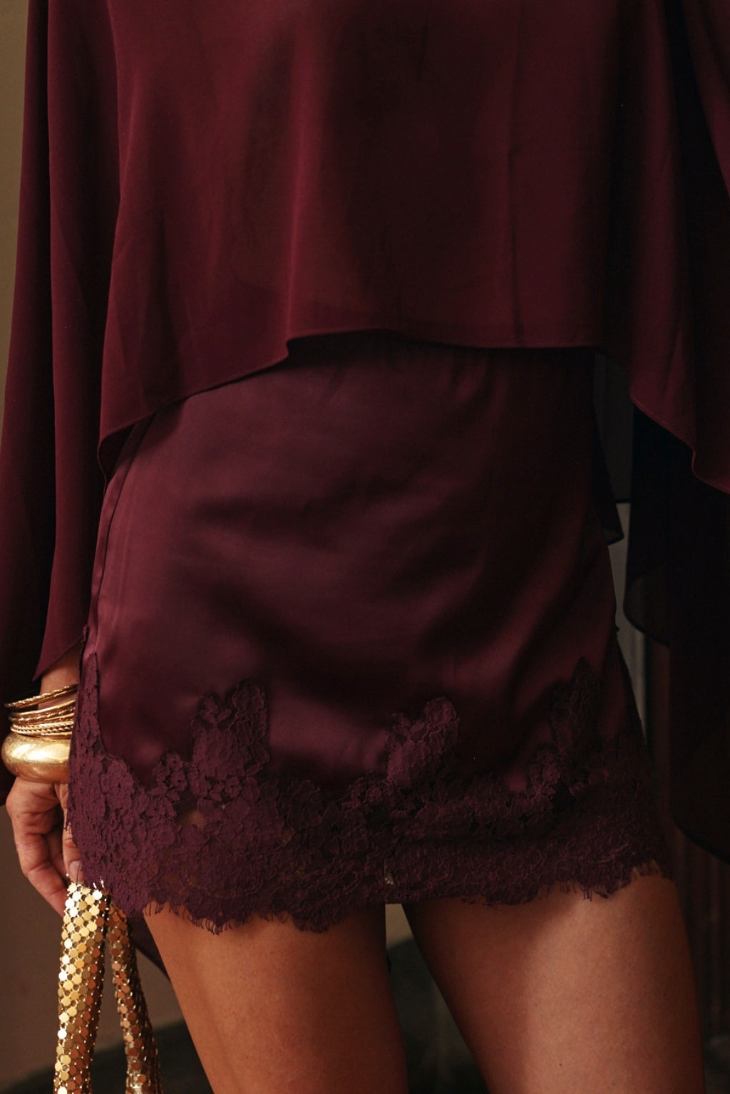 Close-up of a burgundy satin mini skirt with a scalloped lace hem, worn with a matching sheer top, gold bangles, and a gold mesh bag.