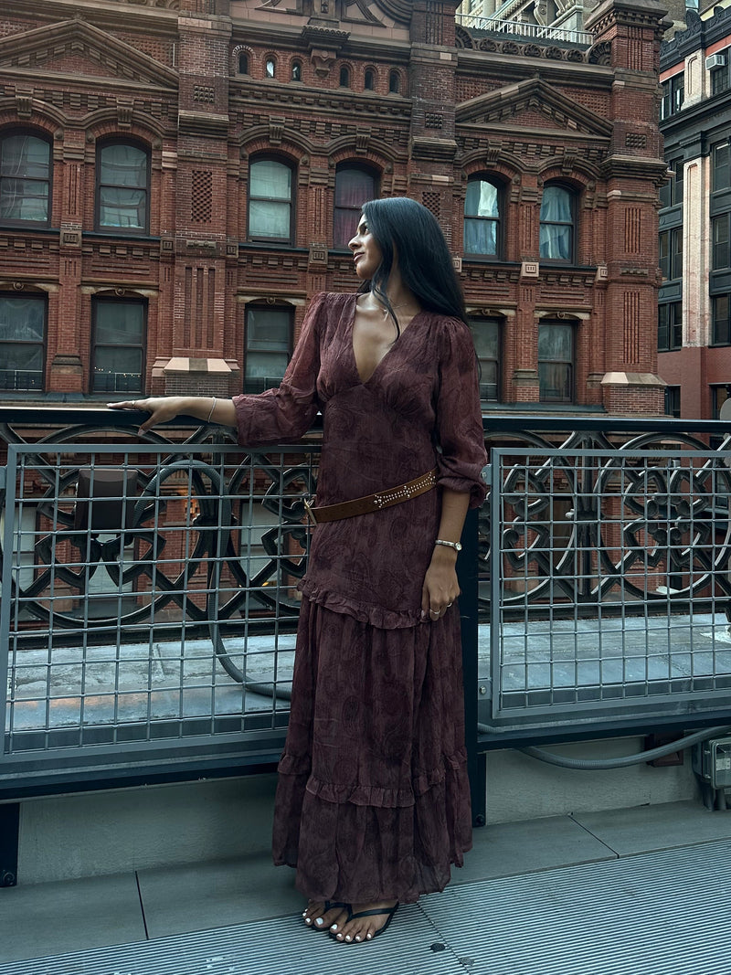 Woman wearing a long-sleeve brown ruffled maxi dress cinched with a braided belt, standing on a rooftop walkway against brick buildings.