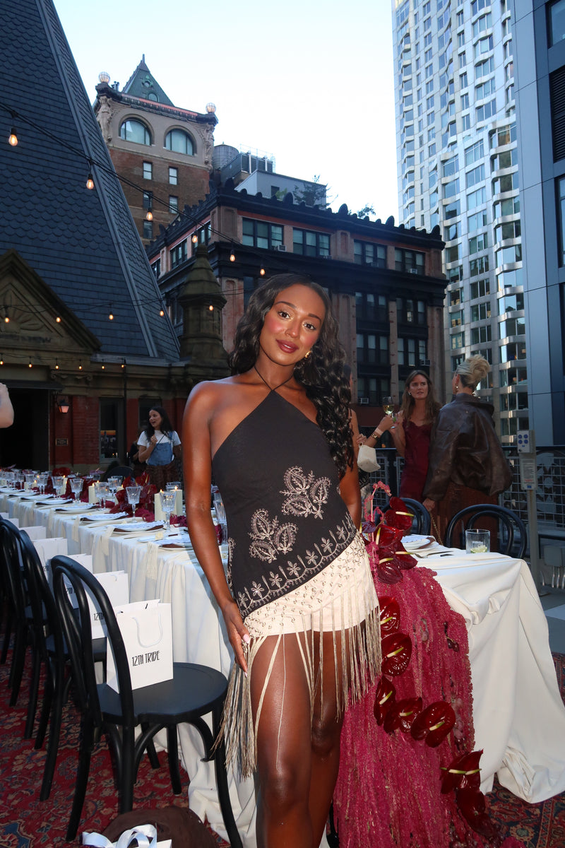 Woman posing at an outdoor rooftop dinner event wearing a black and white fringe dress with butterfly embroidery, city skyline behind her.