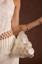 Model wearing the 12th Tribe Adara Ivory Shell Payette Bag, featuring ivory shell embellishments, a gold beaded handle, and a structured base, styled with the Finnian Ivory Jacquard Trousers and Liora Ivory Jacquard Halter Top.