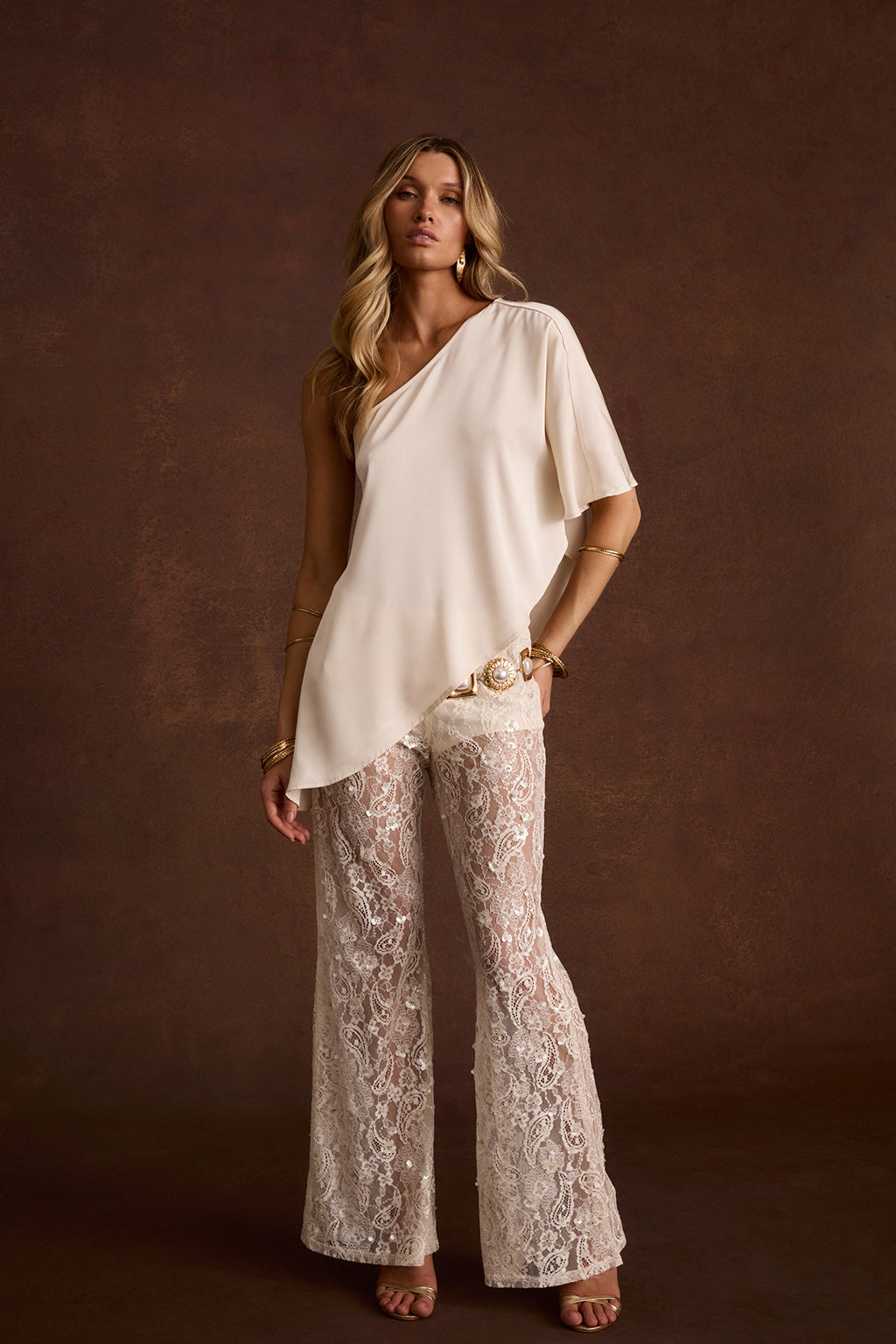 Ivory one-shoulder crepe top and sequin lace paisley flare pants styled with a gold statement belt.