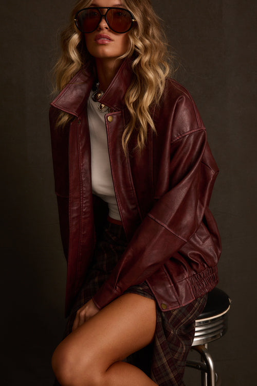 Woman in aviator sunglasses seated on a metal stool wearing an oversized burgundy leather jacket over a white tee and plaid skirt.