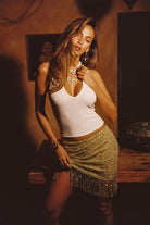 Model wearing the 12th Tribe Luna Cove Sage Fringe Mini Skirt, showcasing a sage green, beaded fringe hem, and sequined detail, styled with the Emily White Ribbed Halter Top, Angelica Gold Pearl Cross Statement Necklace, and Runyon set of 2 cuff bangles.