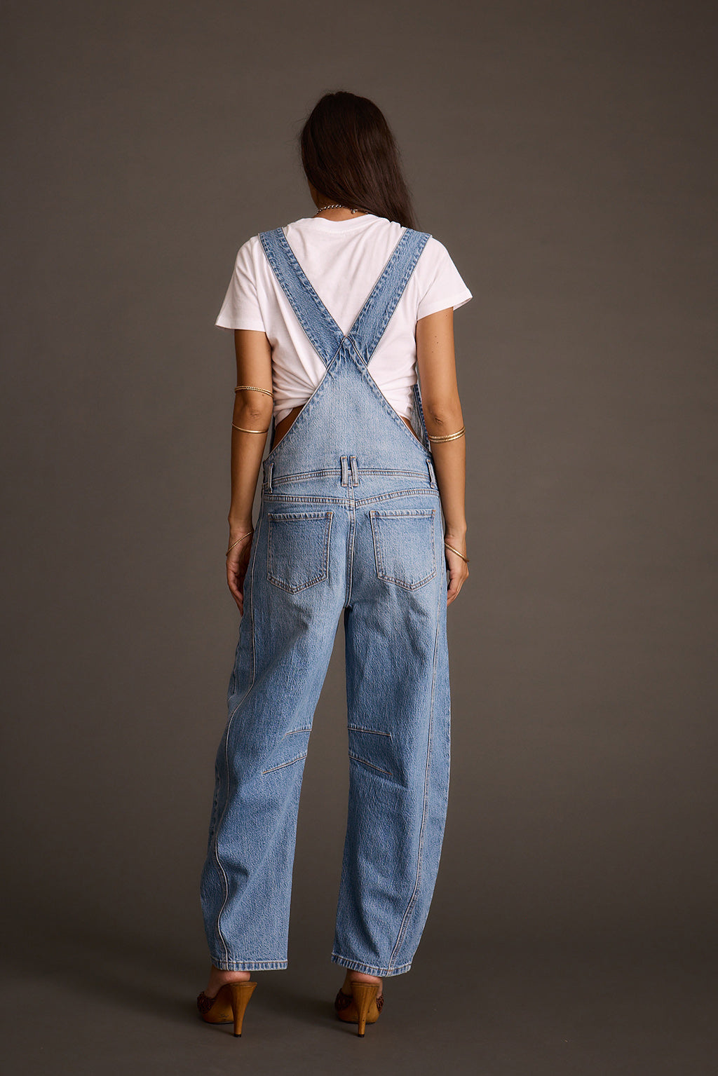 Woman wearing light-wash blue denim barrel-leg overalls, back view