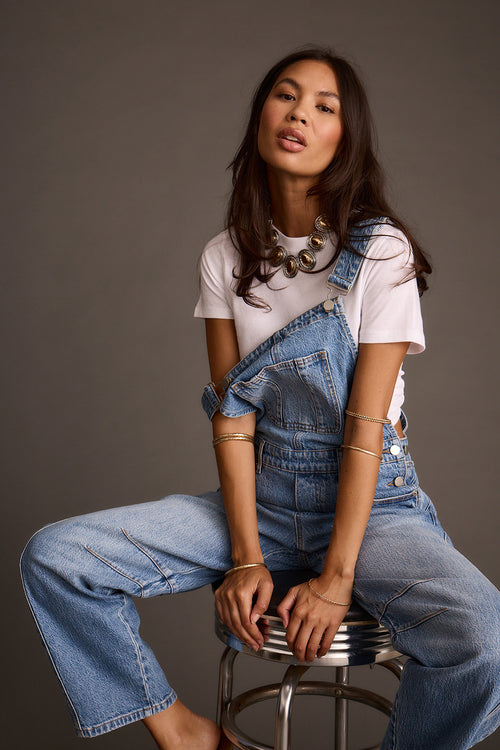 Woman seated wearing light-wash blue denim barrel-leg overalls with one strap off the shoulder