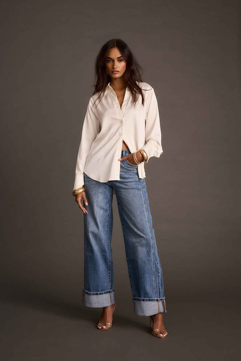 Woman standing in ivory satin button-down shirt tucked into wide-leg jeans with heeled sandals