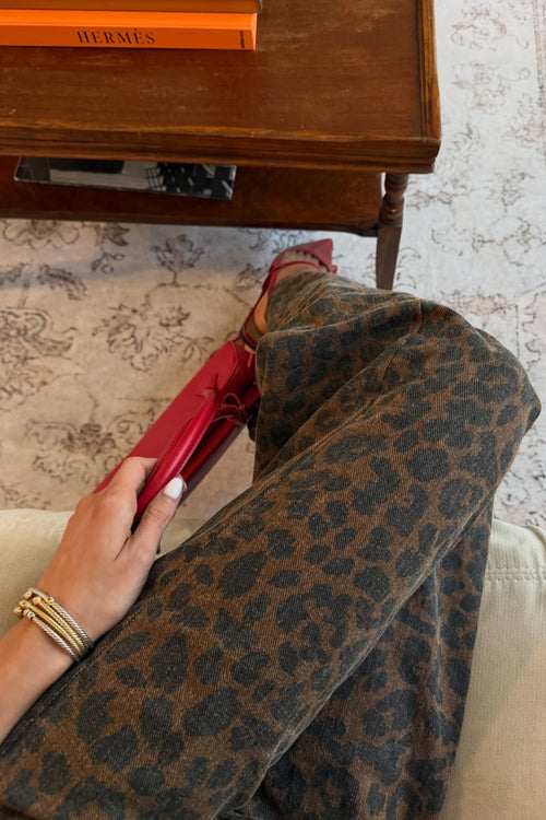 Close-up of brown leopard print denim jeans worn with red pointed heels, with a hand holding a red clutch beside a wooden side table