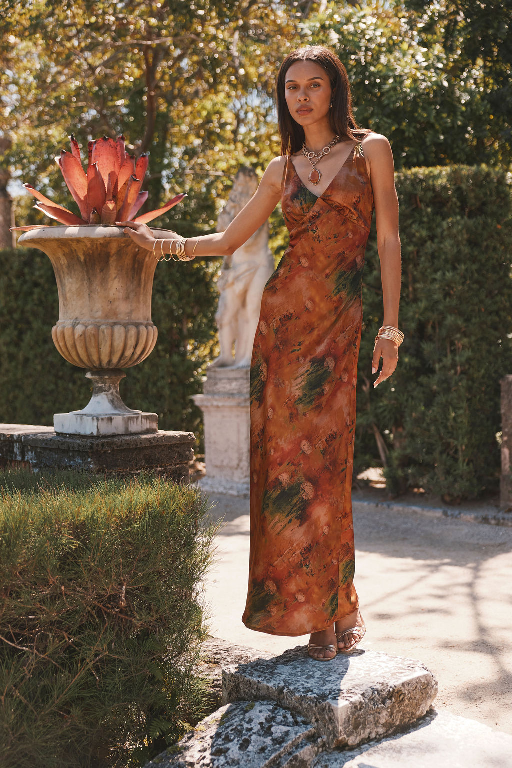 Rust printed satin maxi dress worn beside a stone garden urn, gold stacked bracelets.