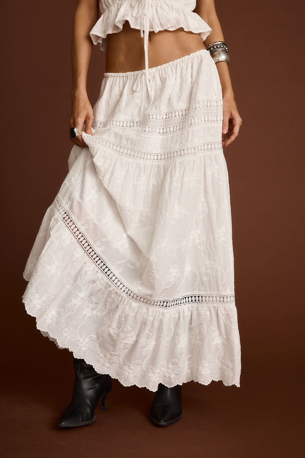 Model wearing the 12th Tribe Nomad White Embroidered Maxi Skirt, a low-rise maxi skirt styled with an adjustable drawcord waist for high, mid, or low-rise fit, featuring delicate floral embroidery throughout, perfect for summer soirées, festival-ready looks, and stylish warm-weather outfits