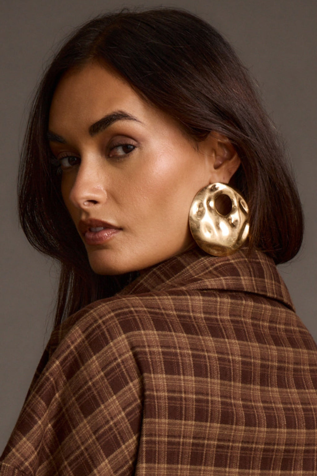 Close-up portrait of a model wearing a chunky gold statement earring with a brown plaid coat
