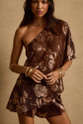 Model wearing the 12th Tribe Anya Mocha Floral One Shoulder Top, a lightweight sheer shimmer one shoulder top featuring an asymmetrical neckline, single sleeve, and asymmetrical high low silhouette in a warm mocha tone. Perfect for warm summer nights, rooftop gatherings, and vacation looks.