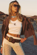 Woman wearing a brown vegan suede fringe jacket with white crop top and cream lace pants against a desert landscape