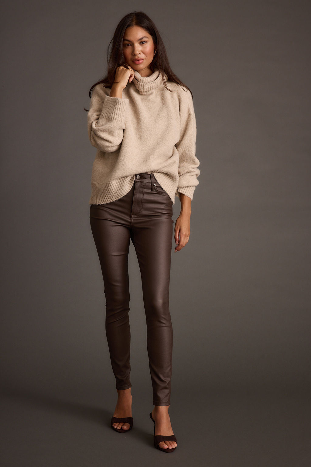 Woman wearing a beige sweater and brown leather pants against a gray background