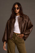 Woman modeling a brown faux-leather jacket over a white top with olive green pants and sunglasses.