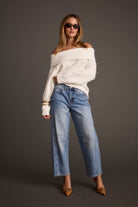 Woman wearing a white off-shoulder sweater and blue jeans against a gray background