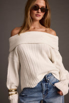 Woman wearing a white off-shoulder sweater and blue jeans against a gray background