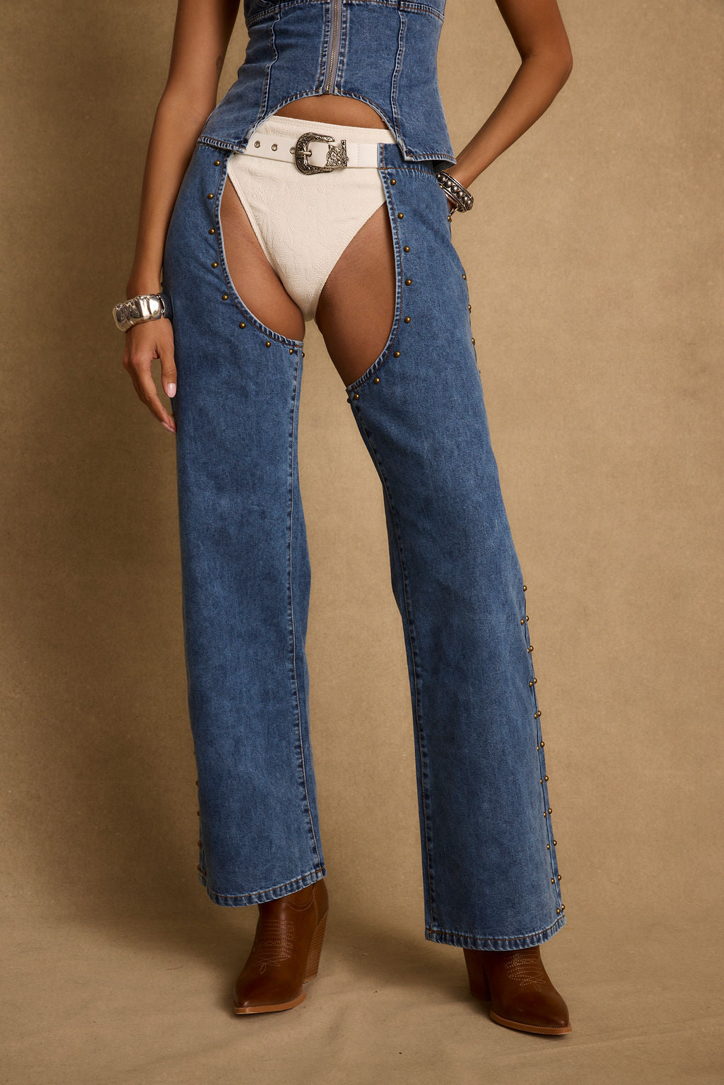 Woman wearing blue denim chaps with bronze studs and cutout legs over white undergarments, a white studded belt with an ornate buckle, and silver cuff bracelets