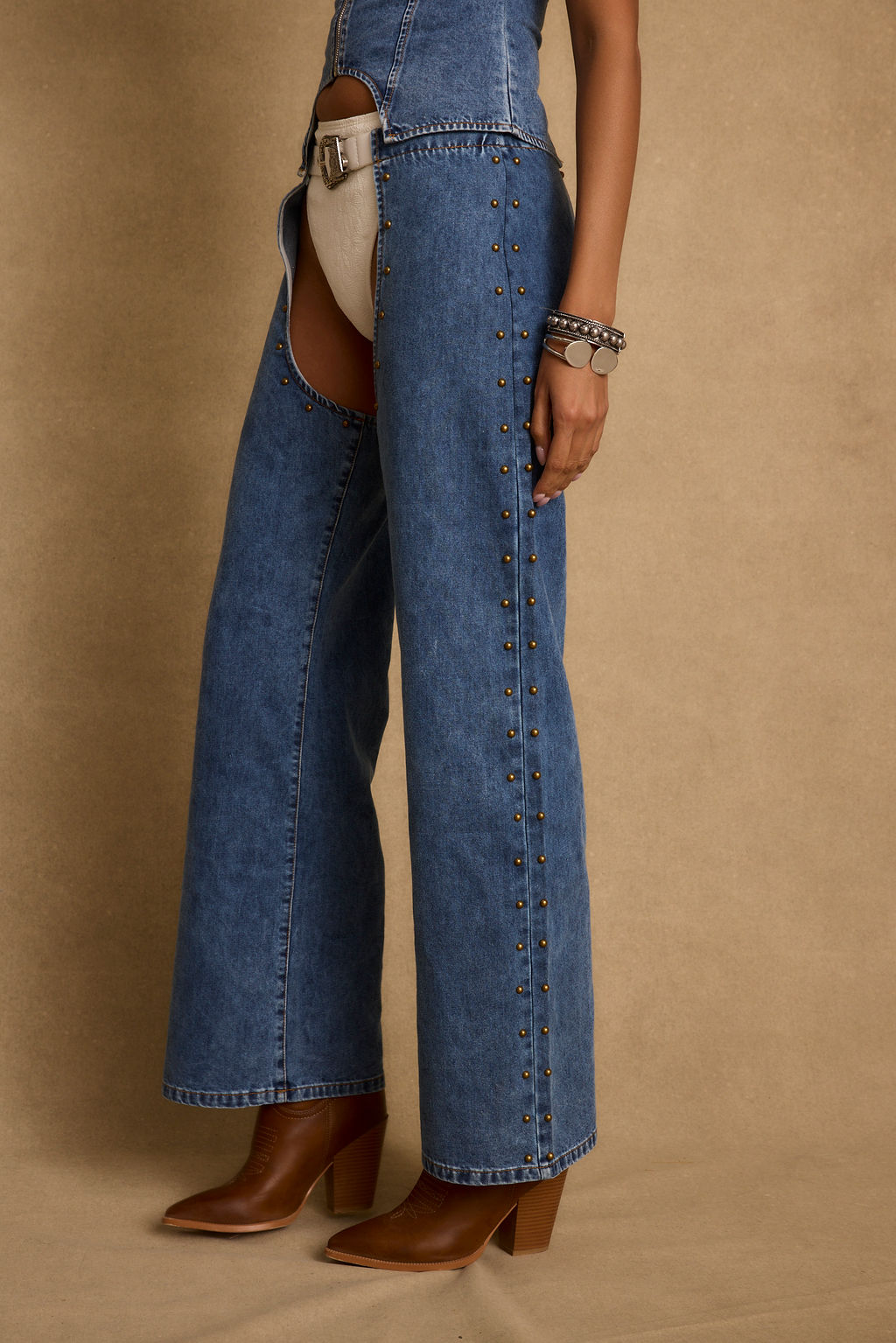 Blue denim chaps with bronze stud detailing worn over a white undergarment with brown leather boots and a silver chain bracelet