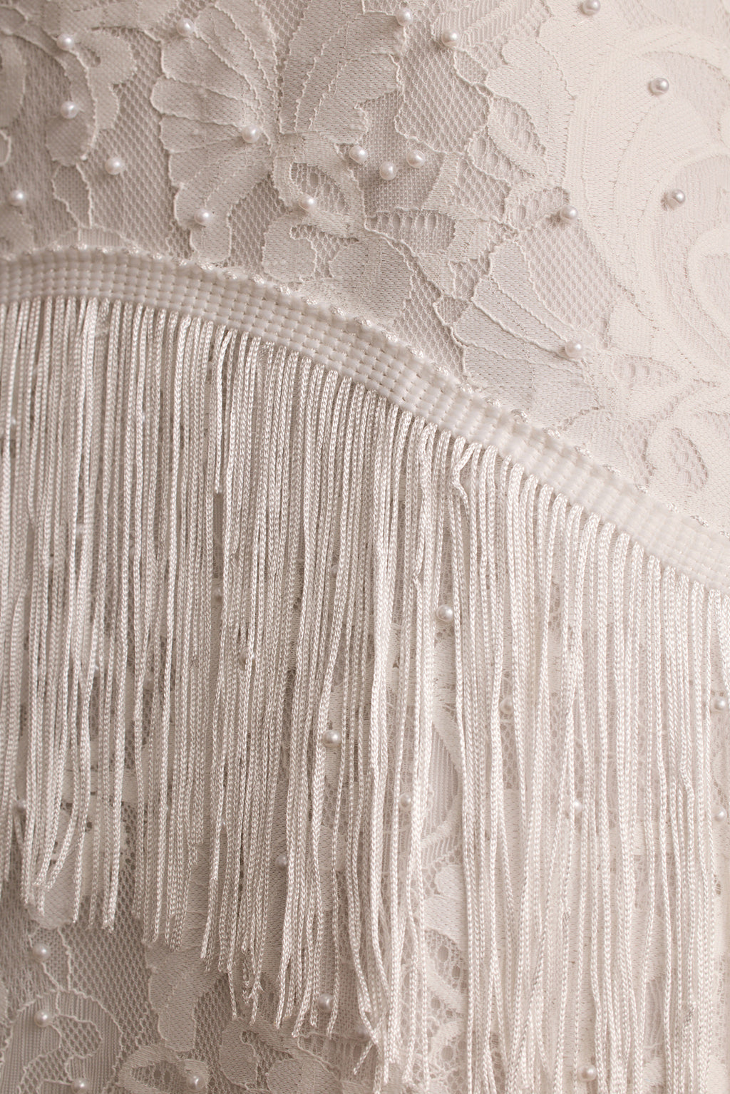 Close-up of white floral lace with pearl beading and silky fringe trim detail.