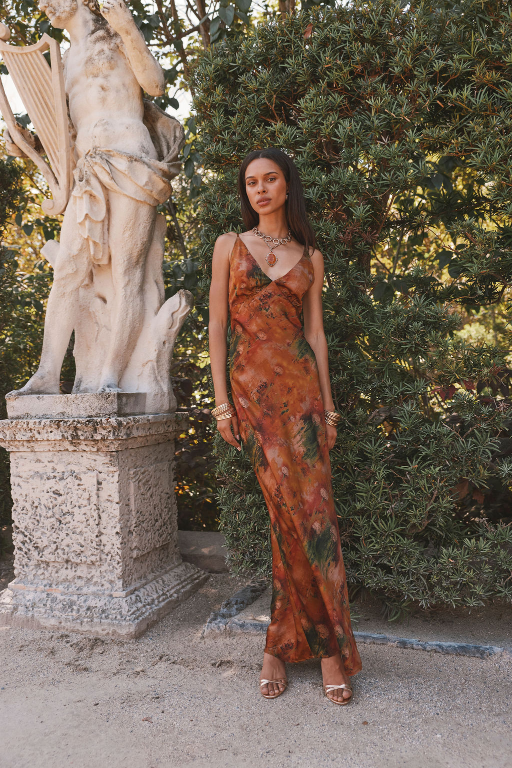 Rust printed satin maxi dress with v-neckline, styled with gold jewelry in a garden.