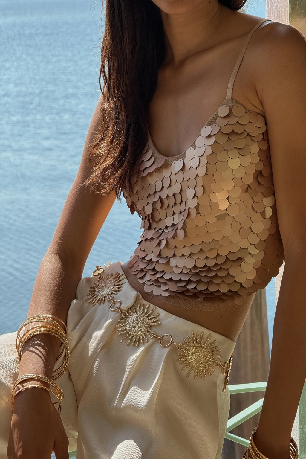 Rose gold payette sequin tank styled with cream trousers and sun-charm belt by the water.