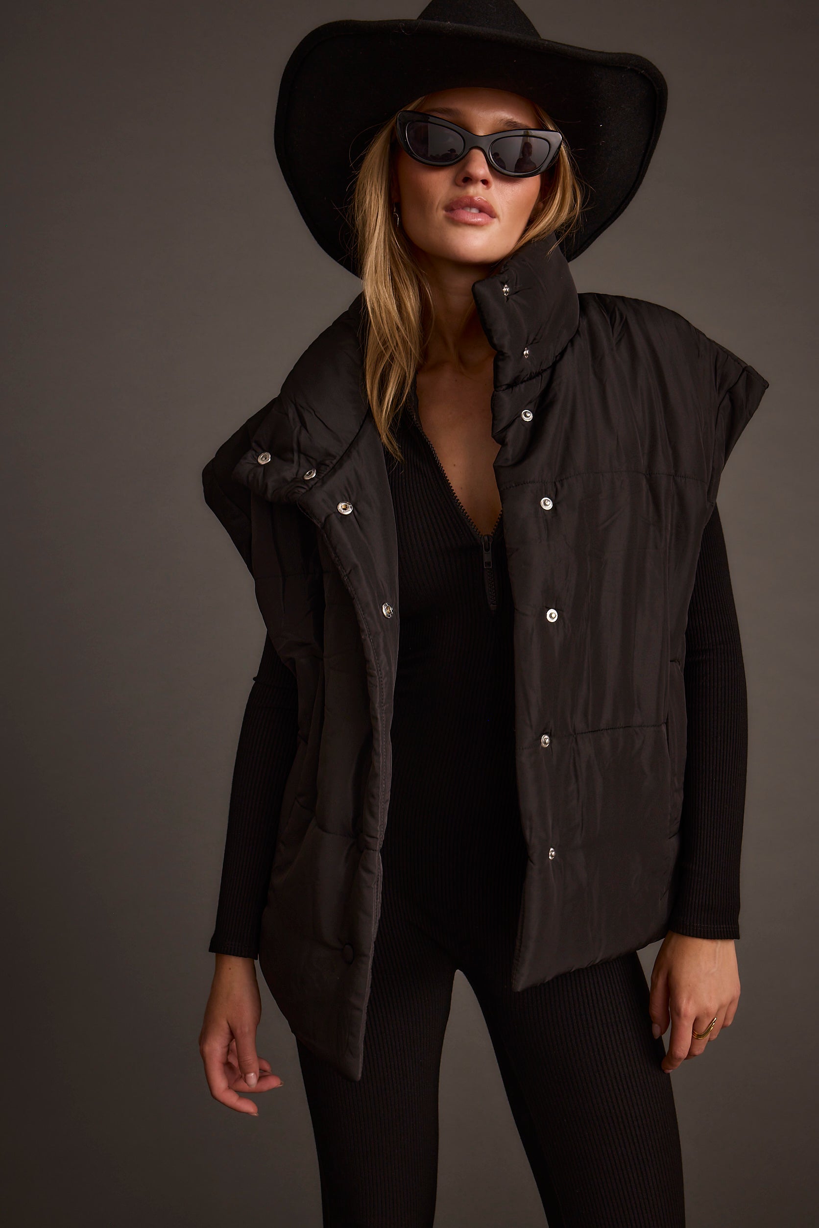 Woman wearing a black outfit with a hat and sunglasses on a dark background