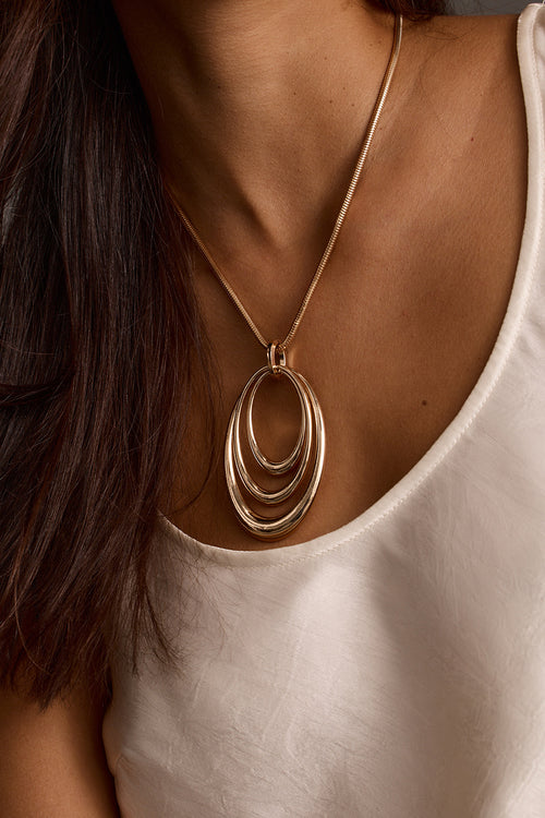 Close-up of a gold pendant necklace with layered oval rings over a white satin top