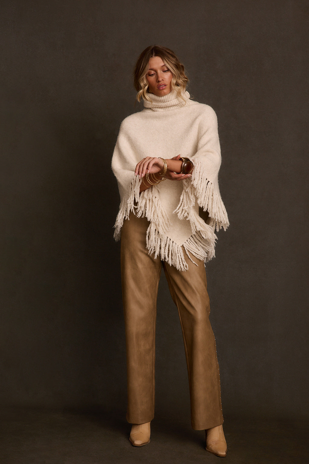 Knit poncho side angle on model with camel pants and gold bangles, fringe detail visible