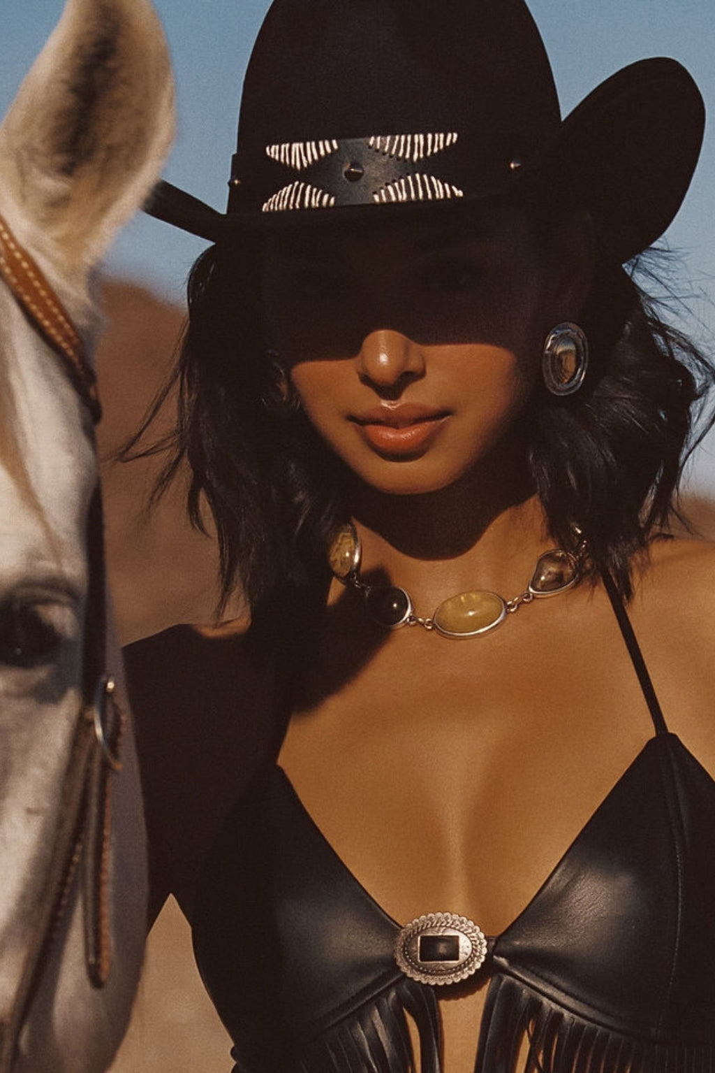 Talon Black Suede Cowboy Hat with zebra-print band detail worn with a black leather fringe top and silver concho jewelry beside a white horse in the desert.
